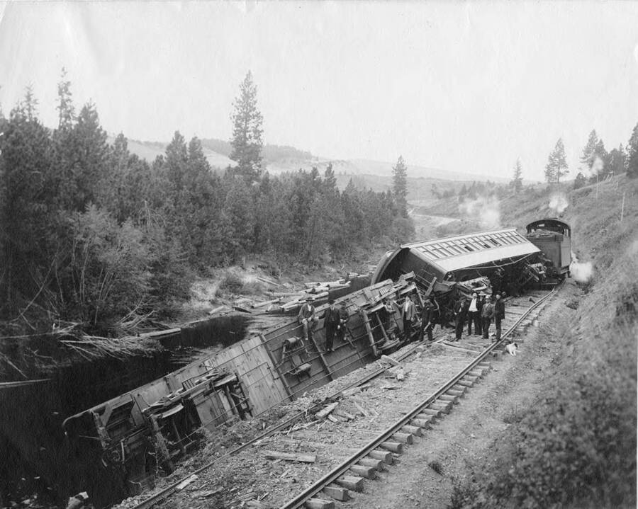 A train wreck  along the Palouse river mainline between Potlatch and Palouse (information taken from back of photograph). Men stand near one of the cars that is over halfway down the embankment while the car behind them tips precariously.