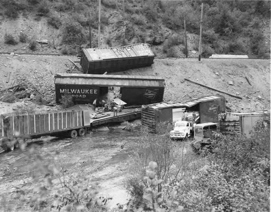 Cars from the Milwaukee Railroad litter the St. Joe river and embankment. Men work in the open cars to insure the safety of the lumber boards in the cars.