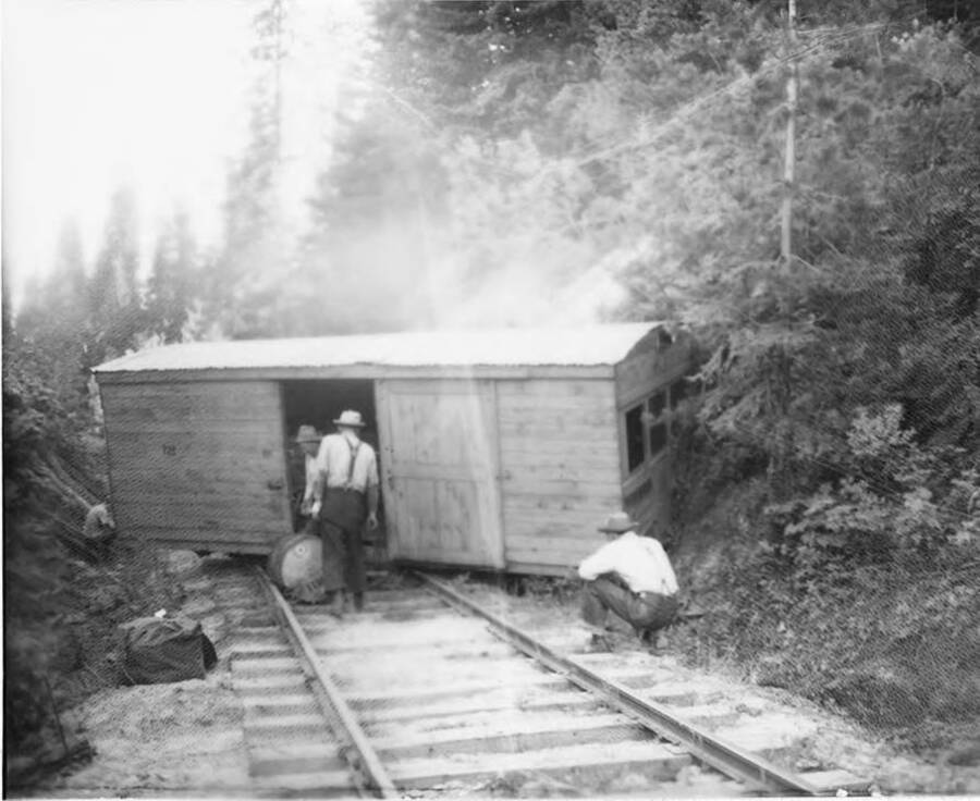 A speeder car is derailed off the tracks. A man kneels to one side of the tracks, while two other men try to reload the car.