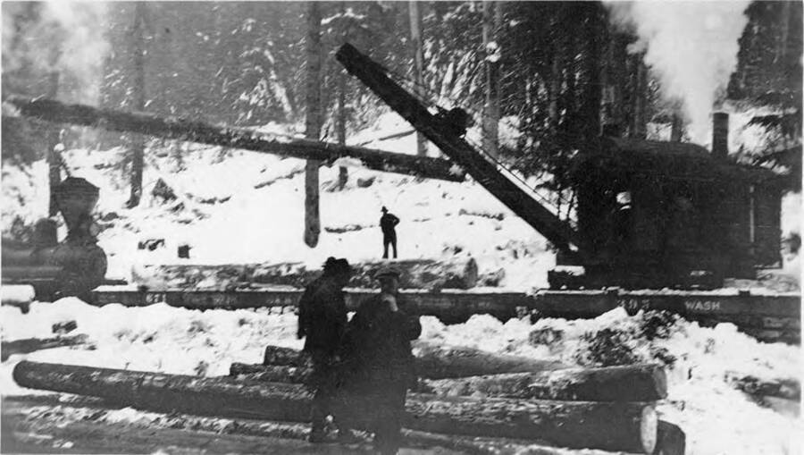Two men stand in front of a crane loading a log onto a flat car.