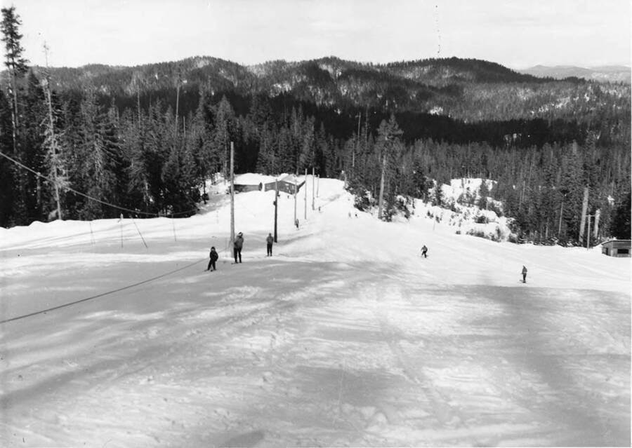 One part of the ski area of Bald Mountain. On the left, skiers use a tow rope to be pulled up the mountain, while on the right, two skiers ski down the hill.
