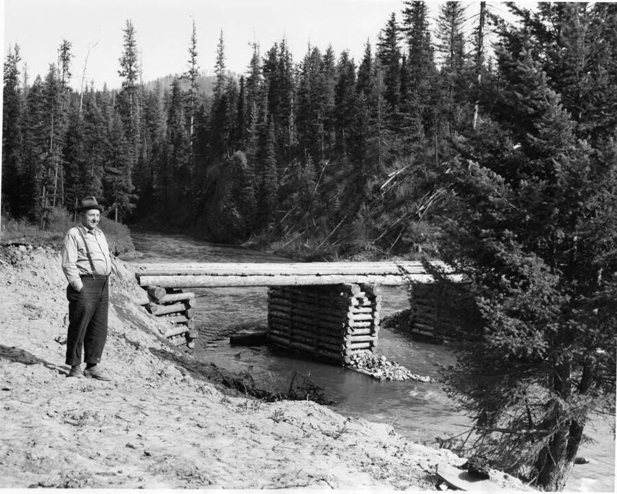 A man stands in front of a bridge that crosses the Potlatch River. According to the description on the back, this bridge is on the way from Helmer, Idaho (which is no Idaho highway 3). The man standing in the picture is Axel Anderson and this bridge is a truck road that replaces the road to camp 4.