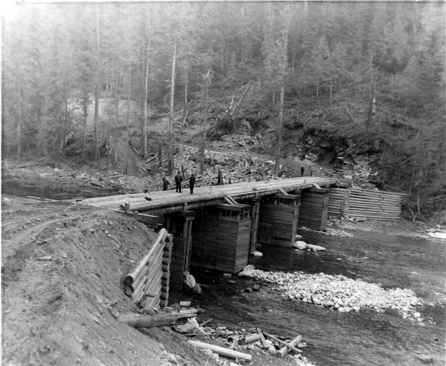 Men stand on a mostly completed bridge. Below them is a creek. On the other side, the road goes up into the forest.