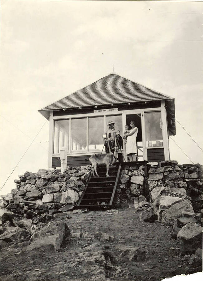 The Iron Mountain lookout station. A man stands with a woman holds a young child at the door. A goat stands on the steps to the cabin.