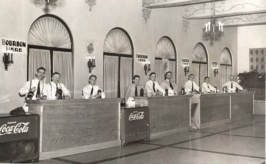 An event for loggers, men stand behind bars, ready to pour drinks. Signs above them read both Bourbon and Scotch here, while between two of the bars are units that have the Coca-Cola name on them. It appears to be at the Lewis Clark Hotel. Listed on the back are the names of the men: JJ O'Connell, C. O. Grave, Lloyd Slongle, Bus Lyells, Leo Bodine, Bob Woods, Steve Summers, unknown, and Ray Hoffman.
