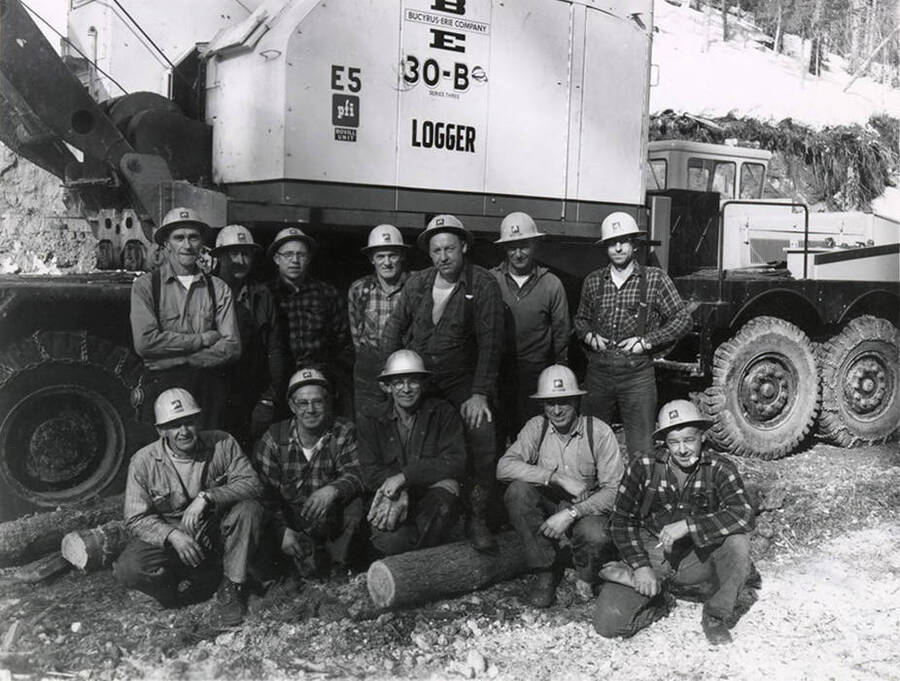 A group of men pose in front of a piece of logging equipment. The envelope holding the photograph says 'personnel - woods. Bovill - CP 43 or 42 crew. Supervisory woods and shop personnel.'