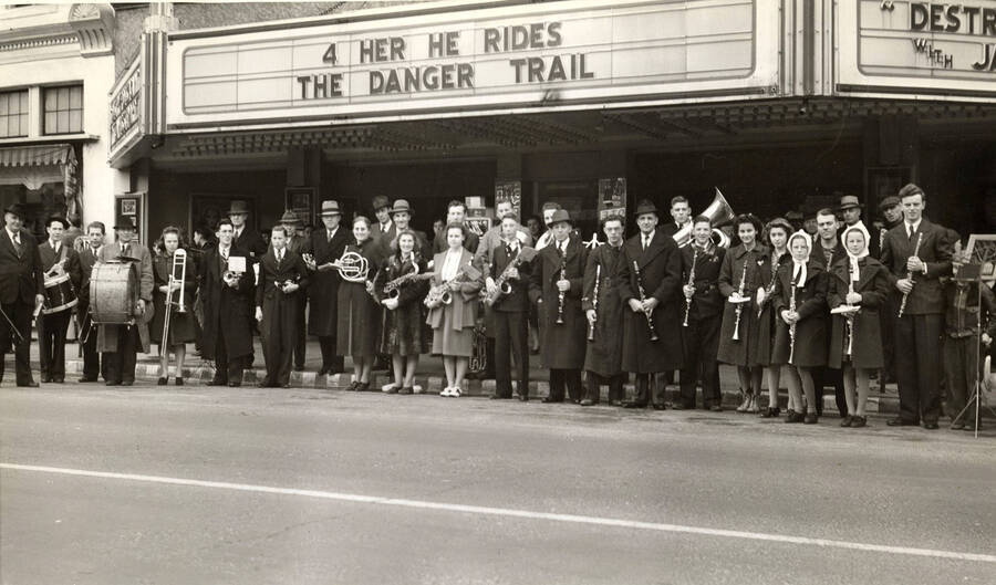 Men and women holding instruments stand in front of the Liberty Theater in Lewiston Idaho. On the marquee above them it reads '4 her he rides' and 'The Danger Trail.'
