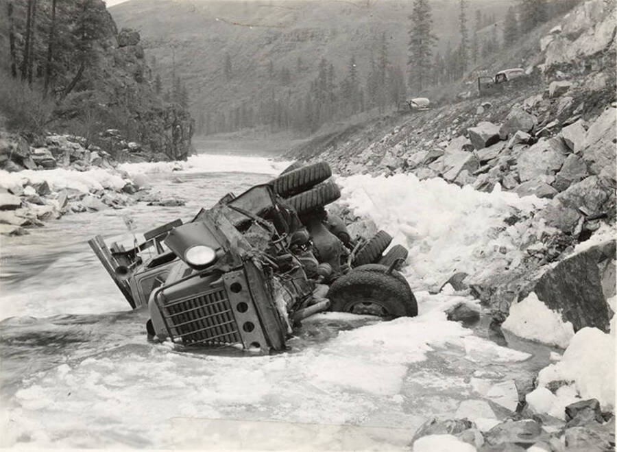 A truck lays on its side in the river surrounded by ice. The description on the back of the photograph reads 'Vehicle Accident. Camp 44 fishhook?'