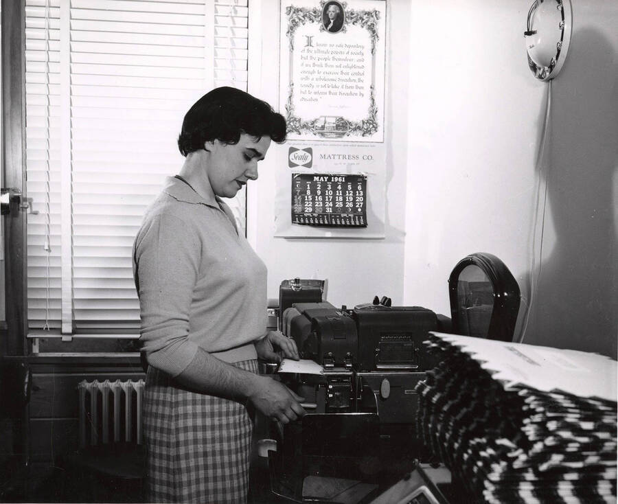 Joan Elsensohn Gruber puts envelopes through a machine. The name of the studio is stamped on the back: 'Imel studio 703 8th St. Lewiston, Idaho.'
