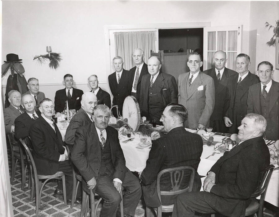 Men sit around a table decorated with wood products. The man in the lower right hand corner is sitting in front of a name tag that reads E. R. Danneman but otherwise the men are unidentified.