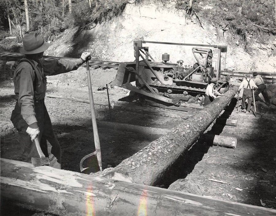 Four men work on a log  next to the railroad tracks. The man closes to the camera holds a logging hook and an ax. On the other end of the log, a man sits in the driving chair of a caterpillar piece of equipment.