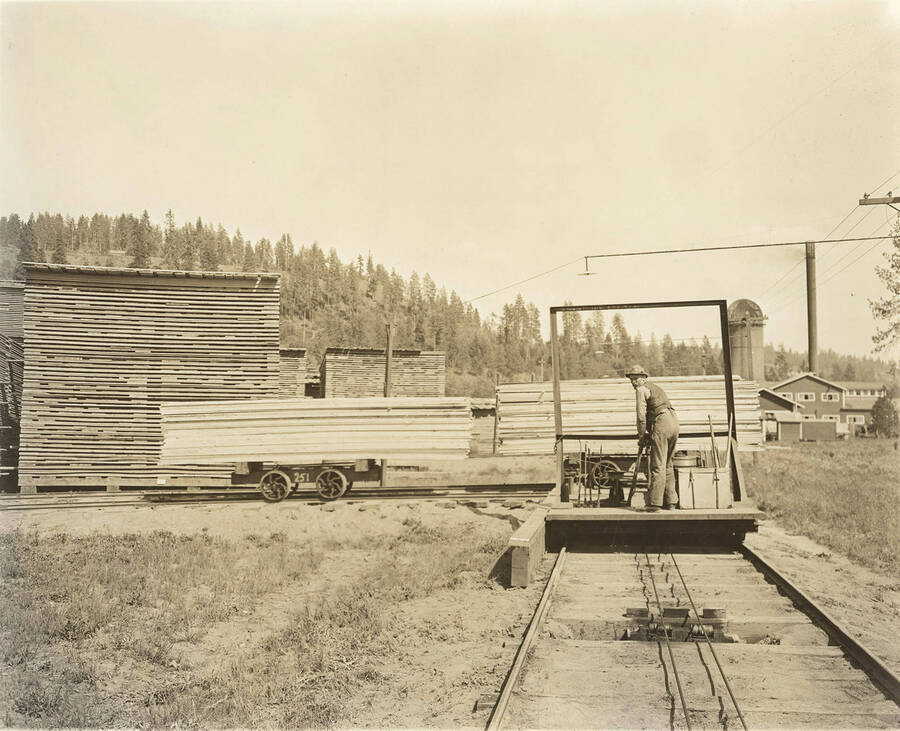 A worker stands on a handcar in the Rutledge Mill lumber yard. Behind him are pallets of lumber and in the distance is the mill.