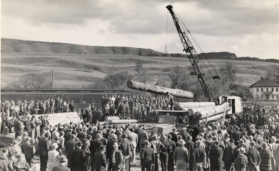 Men and women stand around at the 1940 Lumber Congress, watching a crane lower logs onto the field. Behind the group on the left stands a Northern Pacific railroad car.
