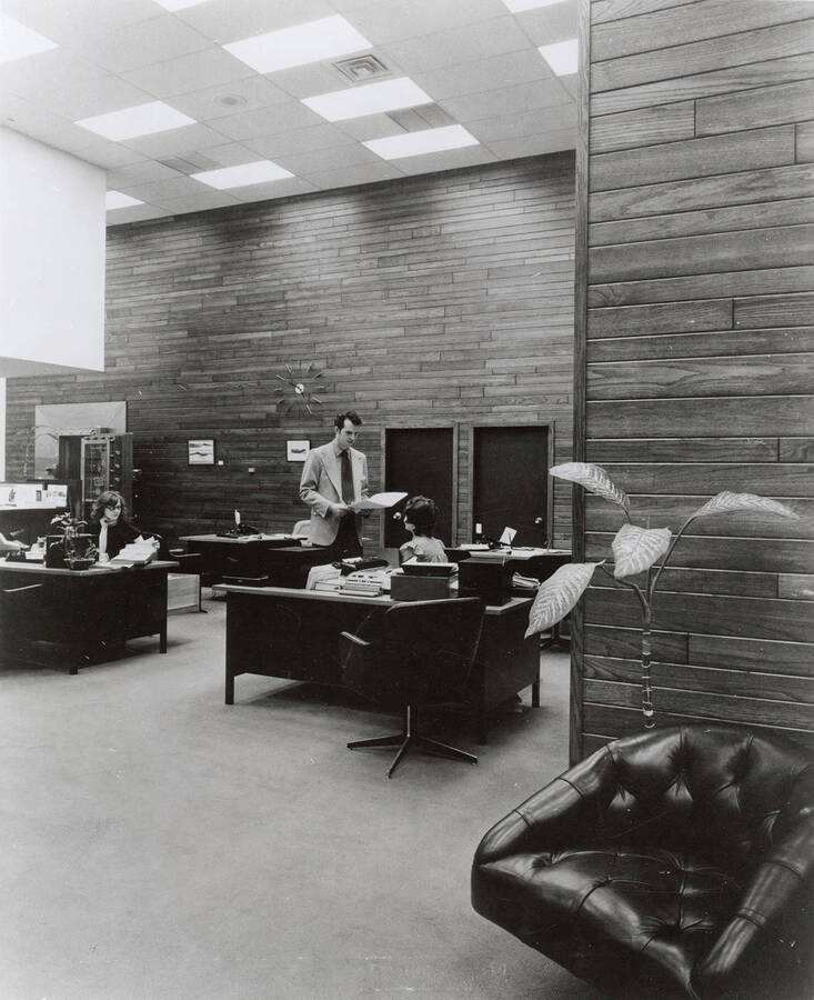 An office showing the walls made of Potlatch Forests, INC. In the center, a man talks to secretary.