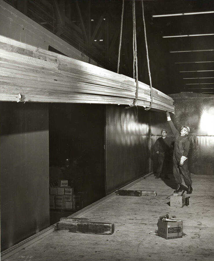 Two men load long boards of lumber into a train car.