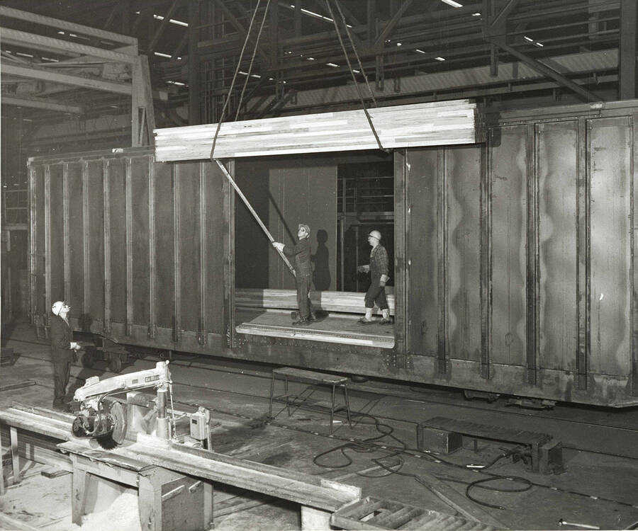 A man uses a tool to help guide the lumber into a railroad car while two men watch.