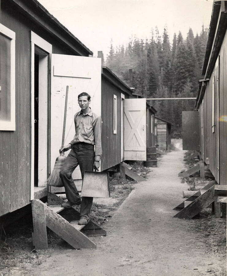 A man stands on the steps to his bunk house with a broom in one hand and a dust pan in the other. All down the row, steps can be seen into each bunkhouse.