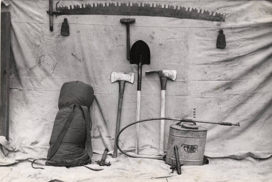 A photograph of many of the tools a lumberjack may use while working in the forest.  At the top is a two-person saw; leaning against the backdrop is a sleeping pack, a shovel, and two types of axes. Also in the kit is some sort of sprayer.