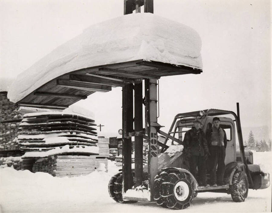 Two men stand on the running board of a forklift that is holding a board covered in what looks to be four or five feet of snow. On the back of the photograph is stamped with Jan. 1952.