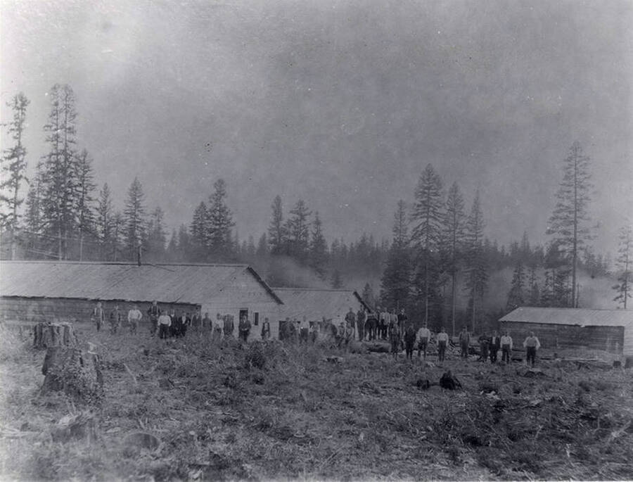 Men pose for a picture in front of buildings at an early logging in Latah county, Idaho. The area in front of the men is cleared of trees while the area behind the buildings is thick with trees.