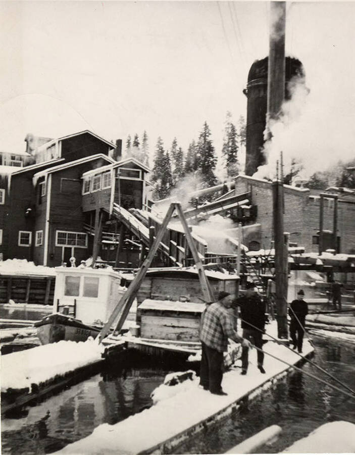 Four men work to pull in a log at the Rutledge Mill in Coeur D'Alene, Idaho. Behind them sits a tugboat and the mill. On the back of the photograph: 'Lyman Acre(?), Robert Matthews, James  Roe, and John Doucey (?).