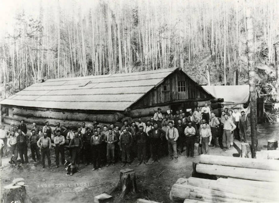A group of loggers (and a dog) stand in front of building  in an unknown logging camp near Bovill, Idaho. Behind the men and buildings are un-harvested trees, while in the lower right hand section of the photograph are harvested logs.