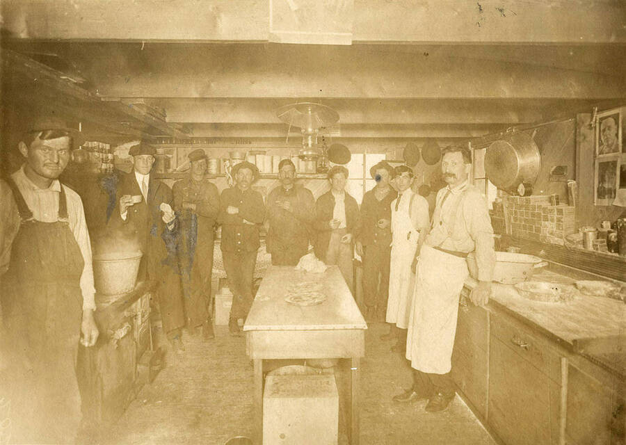 Men stand in an early logging camp kitchen. On the left is the stove, in the middle a island table, and to the right is another cooking area. Overhead is a lamplight.