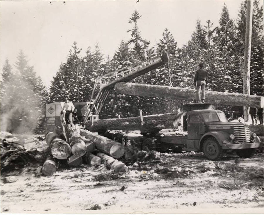 A crane loads a log on to a truck as a man stands on top of the upper arm of the bed of the track.