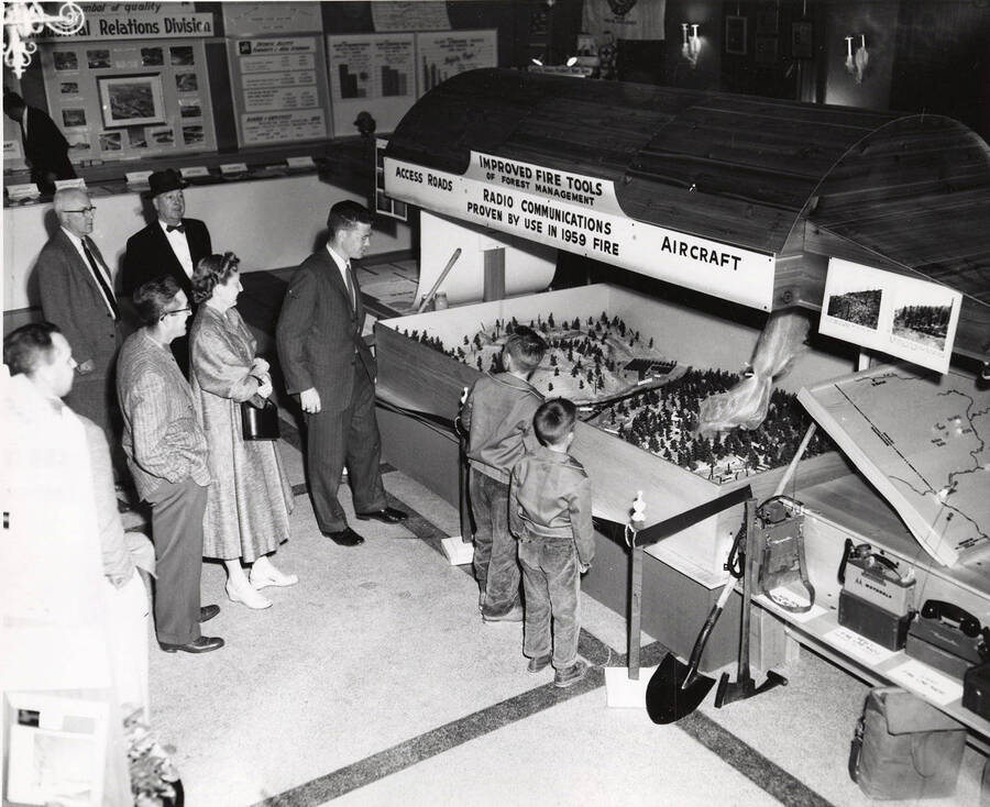 Men, women, and children stand in front of a model displaying the "Improved Fire Tools" of forest management. The model includes three different areas: access roads, radio communications, and aircraft. Off to the right hand side of the model some tools of forest firefighters and communication devices.