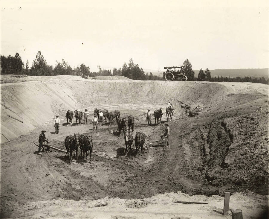 Men stand with teams of horses in a pit. At the rim of the pit in back is an early model of a steamroller.