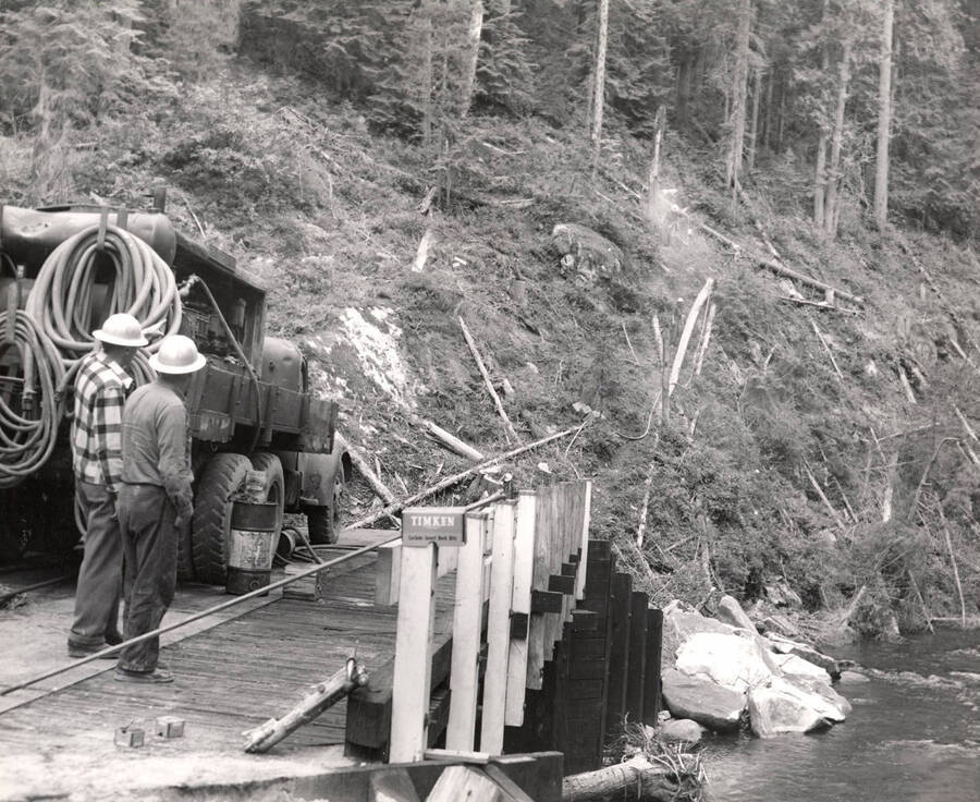 Two men stand on a bridge watching as several other men clear trees and brush from a hillside.