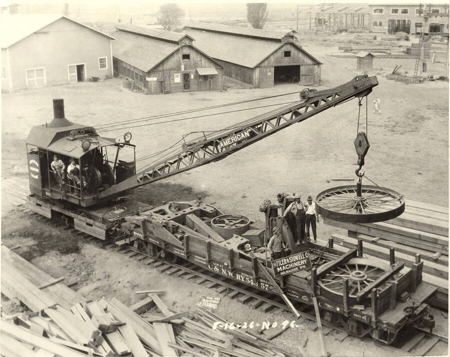 Men stand on a train flatcar as a crane moves a large wheel off. There is another wheel on the flatcar waiting to be moved. In front of one of the men watching the wheel reads a sign 'The Filer & Stowell Co . Machinery. Milwaukee, Wis.'