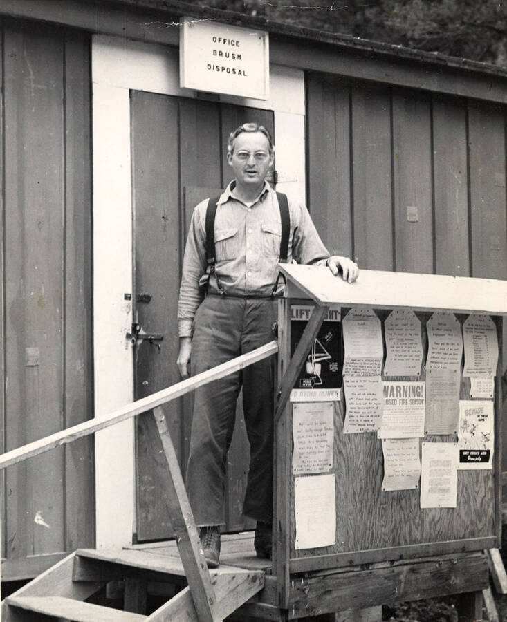 A man stands in front of the office of probably a lumber camp. The sign above him reads 'Office brush disposal'. In front of him is a bulletin board with several notices and pieces of paper.