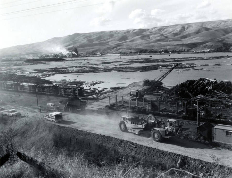 The log pond. Railcars full of logs sit on the left hand side of the photograph waiting to unload while empty ones are on the right hand of the photograph. Heavy moving equipment sits near the empty railcars. A dump truck full of dirt is turning down a dirt road toward the pond and a crane.