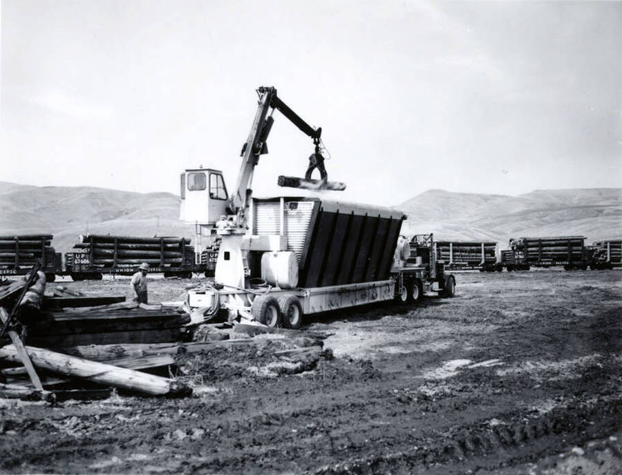 A man watches as a log is lowered into a "smokeless trash burner." Description taken from envelope holding the photographs. In the background are railcars full of logs wait.