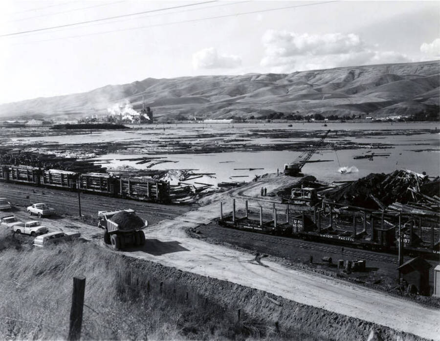 A dump truck carrying dirt sits on a road next to the log pond. Also in the picture, railcars sit full of logs waiting to deposit them in the pond. On a strip of land jutting into the pond, a crane's equipment works in the pond. Empty railcars are on the right-hand side of the picture.