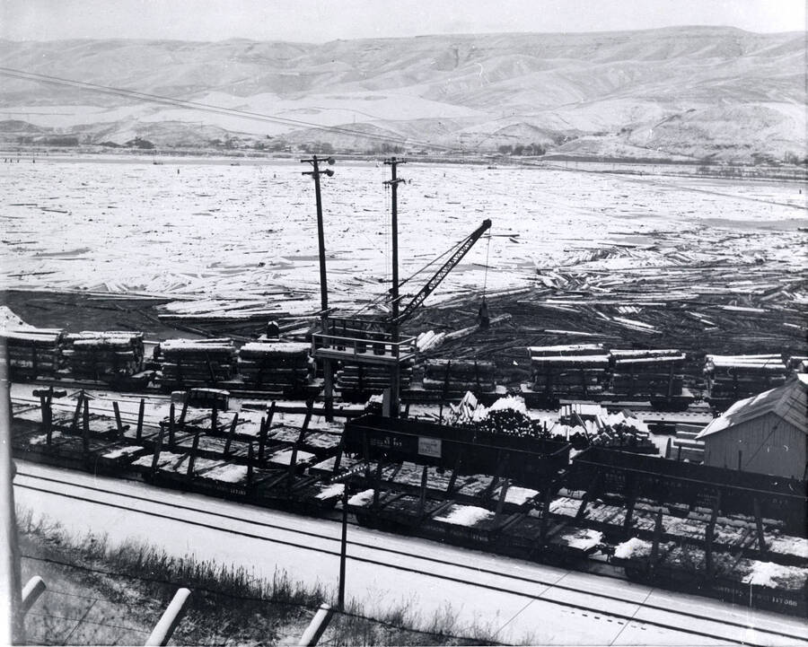 In the foreground, snow-covered railcars sit empty while in the center of the picture, a crane works to remove logs from loaded railcars and put them into the log pond. In the background, snow covered logs sit in the pond. Date on the back of the photograph says December 1959.