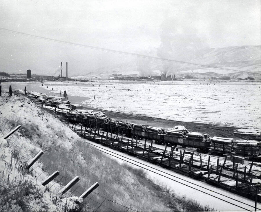 Empty snow-covered flat railcars sit next to full railcars. Behind them lays the log pond which is covered in snow. In the background of the photograph is the sawmill. The back of the photograph says December 1959.
