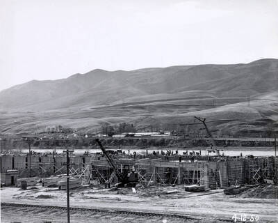 Construction of one of the mill buildings. A crane is at the center of the photograph with two men standing underneath it. The date written on the photograph is 4/12/1950.