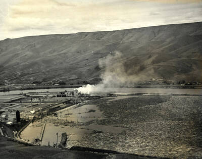 An overview photograph of the log pond and sawmill looking north. In the background, runs the Clearwater river.