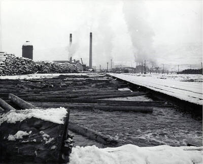 Logs float in choppy water in the Clearwater Mill log pond. At the back of the photograph, snow covered logs lay stacked. On the back of the photograph it lists December 1959 as the date.