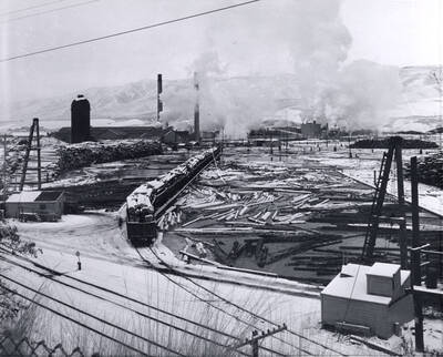 The log pond covered in snow. Loaded railcars sit waiting to unloaded into the log pond are covered with snow. December 1959 is written on the back of the photograph.
