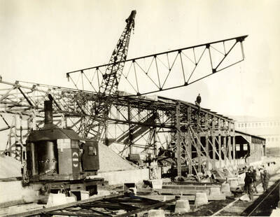 A locomotive crane lifts a piece of framing. Two men sit atop the framing as several men watch below.