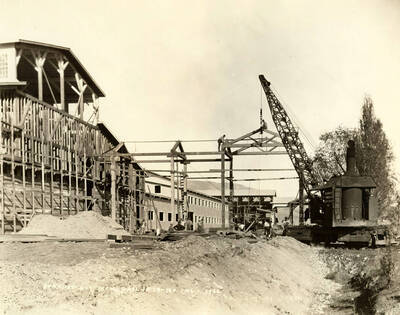 A crane works to help build the roof supports. A man sits atop the roof support near the crane while another stands on a different part of the framing. Several men work below. Written on the picture is 'Stacker Building and Mon-Rail 10/18/1926 No. 146.'