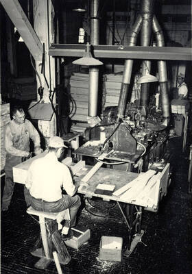 One man is putting the piece of wood into the machine while another ties off sets of the pieces.