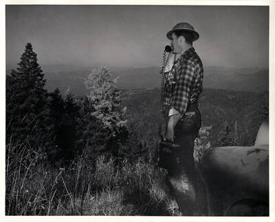 The description on the back of the photograph reads 'Bob Tondevold surveys typical country from atop Bald Mountain. This is terrain which offers 'radar shows' and potential invasion path to enemy. Tondevold uses hand-carried battery-operated transmitter with which he can call in aircraft spotting information.' The writing on the transmitter says 'Littlefone'