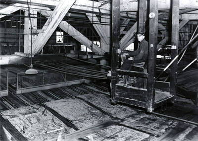 A man sits in a chair over sawdust and scraps of wood while holding on to machinery. The description on the back of the photograph says "trimmer man."