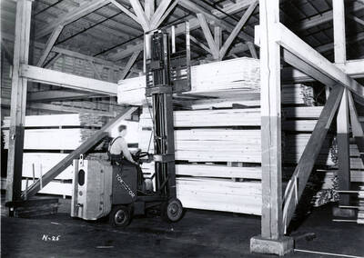 A man uses a forklift to lift a stack of lumber on to a rack. The description on the back reads "Lumber carrier, #24 pressed shed."