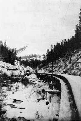 A timber flume on along Tom Taha Creek by Glenwood Road. Haystack Mountain is visible in the background. Next to the flume stand telephone poles and lines.
