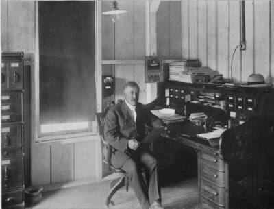 William Deary sits in the office in Potlatch. Scanned from the book 'Potlatch Lumber Company: Manufacturers of Fine Lumber.' The back cover says 'Photographed and published by F. D. Straffin, 336 Riverside Ave. Spokane Wash. Printed by Inland Printing Co. Spokane, Wash.'